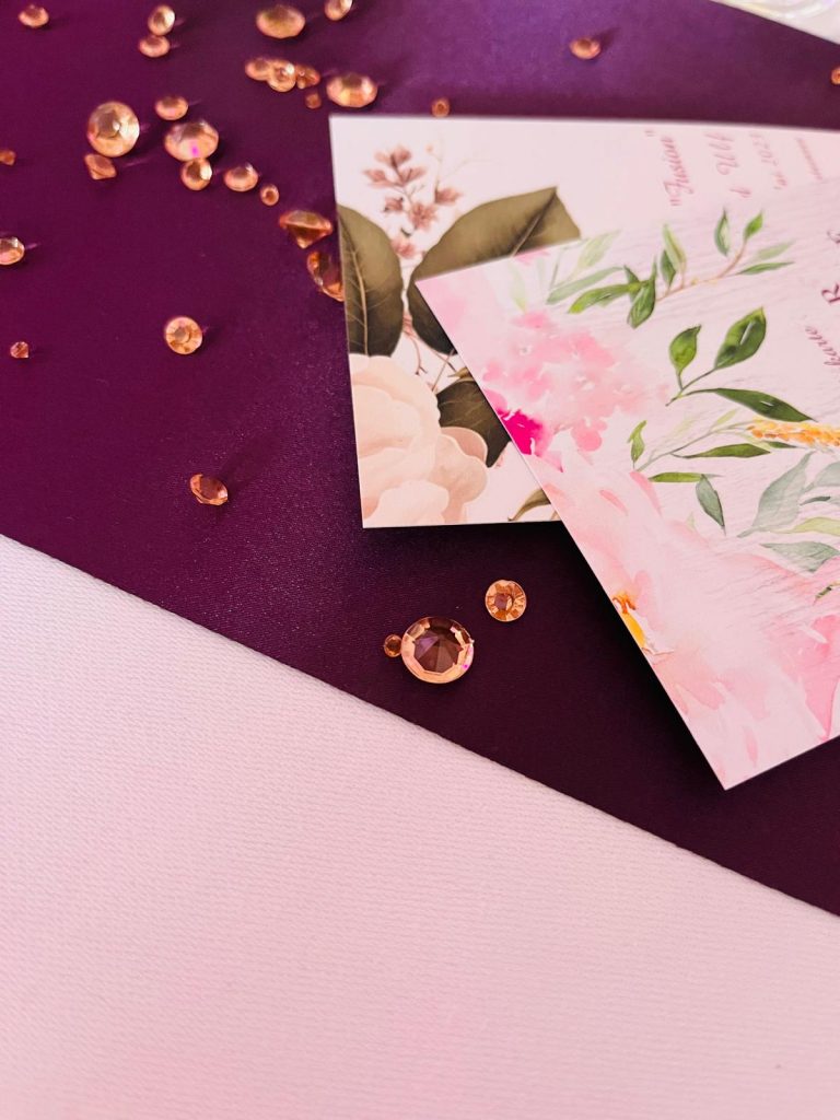 Hochzeitseinladungen mit einem romantischen, floralen Aquarell-Design liegen auf einem dunkelroten Umschlag und sind mit funkelnden, roségoldenen Schmucksteinen dekoriert.