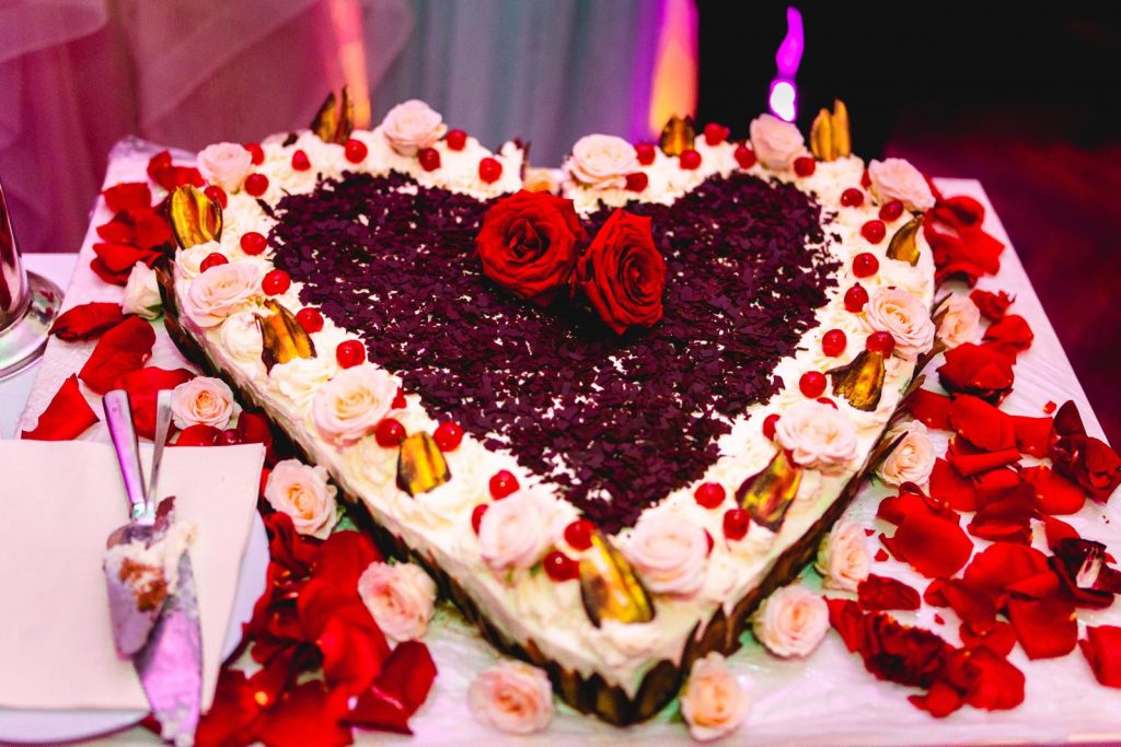 Eine große, herzförmige Hochzeitstorte, vermutlich eine Schwarzwälder Kirschtorte, die mit Sahne, Schokoraspeln, Kirschen und echten roten und weißen Rosen aufwendig dekoriert ist.